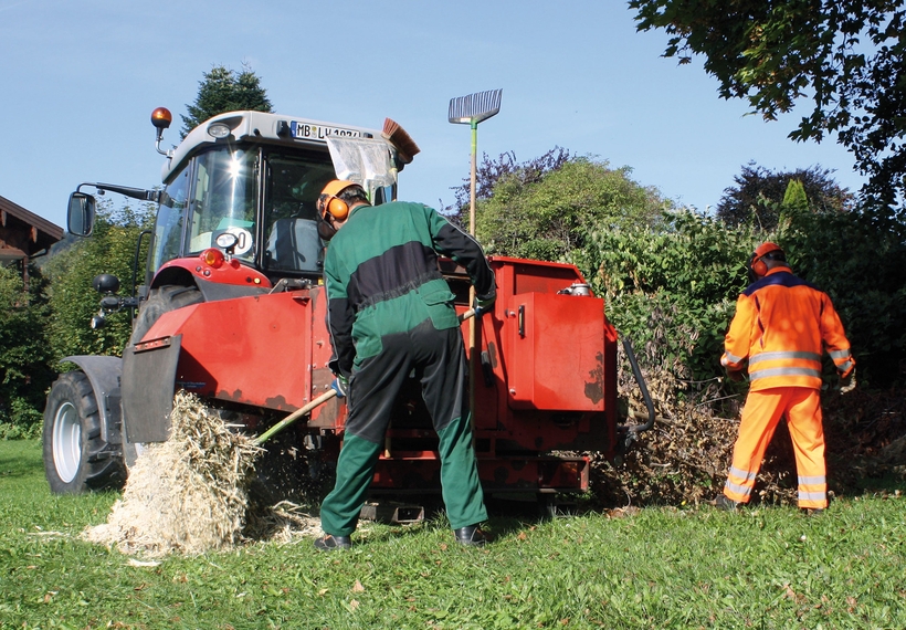 Traktor mit angeschlossenem Holzhäcksler; zwei Arbeiter in Schutzkleidung und Helmen (grün, orange) füttern Äste; Hackschnitzel fallen auf Gras; Bäume und Häuser im Hintergrund