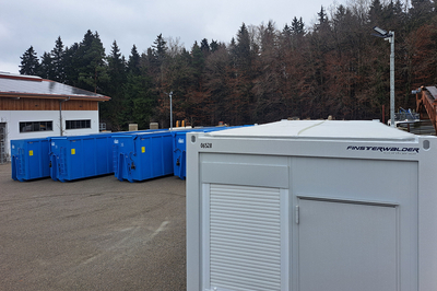 Auf dem Bild sind mehrere blaue Container in einem Außenbereich zu sehen, umgeben von Bäumen und einem Gebäude.