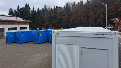 Auf dem Bild sind mehrere blaue Container in einem Außenbereich zu sehen, umgeben von Bäumen und einem Gebäude.