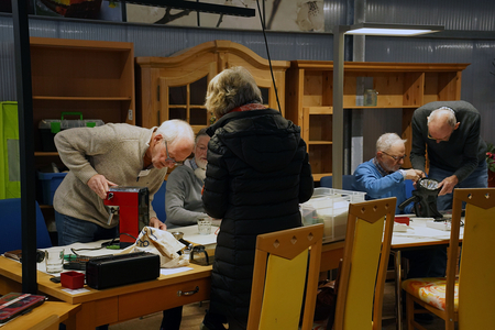 Auf dem Bild sind mehrere älteren Menschen zu sehen, die an Tischen verschiedene Gegenstände reparieren oder untersuchen.