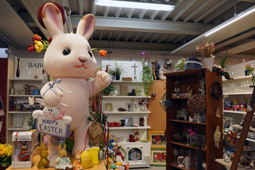 Das Bild zeigt einen großen, niedlichen, plüschigen Hasen, der mit Frühlingsblumen geschmückt ist, in einem Raum voller Deko- und Haushaltsgegenstände.