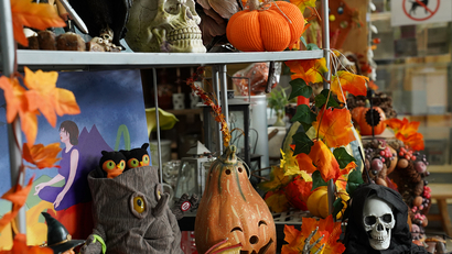 Auf dem Bild sind verschiedene herbstliche und Halloween-Dekorationen wie Kürbisse, eine Totenkopfstatue und bunte Blätter zu sehen.