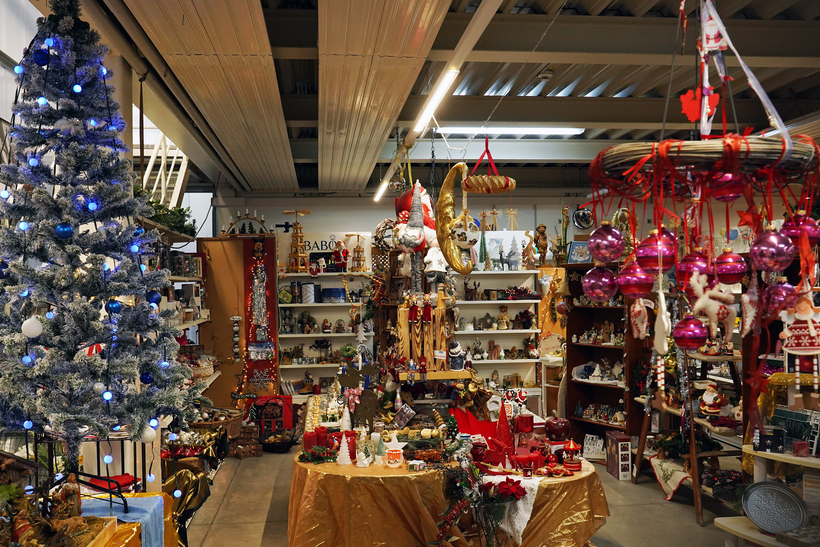 Innenaufnahme eines Weihnachtsdekorationsladens; geschmückter Weihnachtsbaum links; Tische und Regale voller Christbaumschmuck und Figuren; rote und rosa Kugeln rechts; warme Beleuchtung