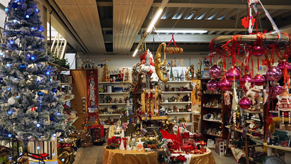 Innenaufnahme eines Weihnachtsdekorationsladens; geschmückter Weihnachtsbaum links; Tische und Regale voller Christbaumschmuck und Figuren; rote und rosa Kugeln rechts; warme Beleuchtung
